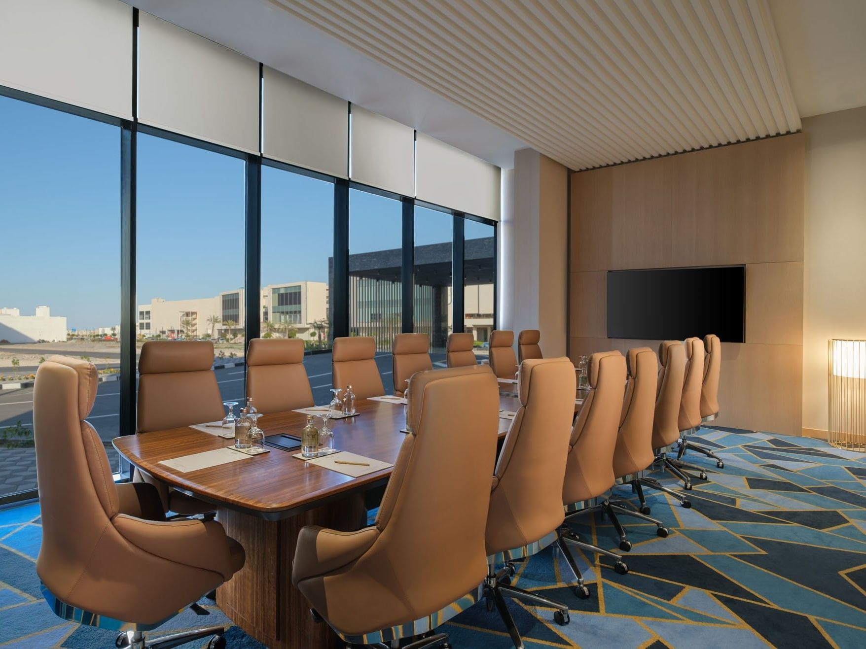 Modern conference room with large windows, a long table, and beige chairs, featuring a flat-screen TV and geometric carpet.