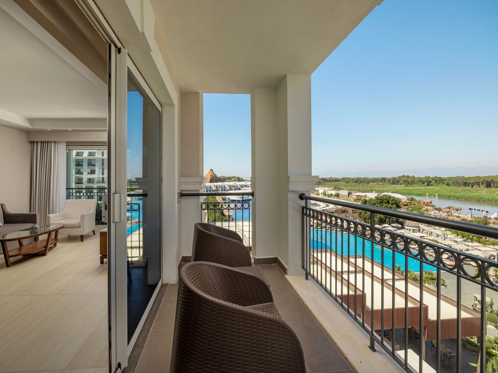Private balcony with wicker seating overlooking a blue pool, lush golf course, and distant landscape at Titanic Deluxe Golf Belek