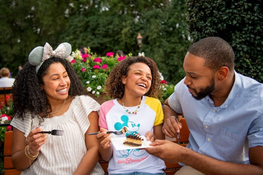 A laughing girl in a Mickey shirt sits between a woman in Minnie ears and a man in a blue shirt, all holding forks and sharing a slice of cake. 