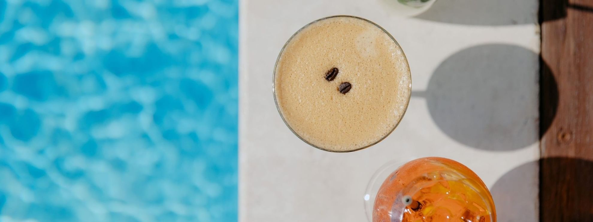 Three drinks by a pool, one with coffee beans, one with mint, and one with an orange slice.