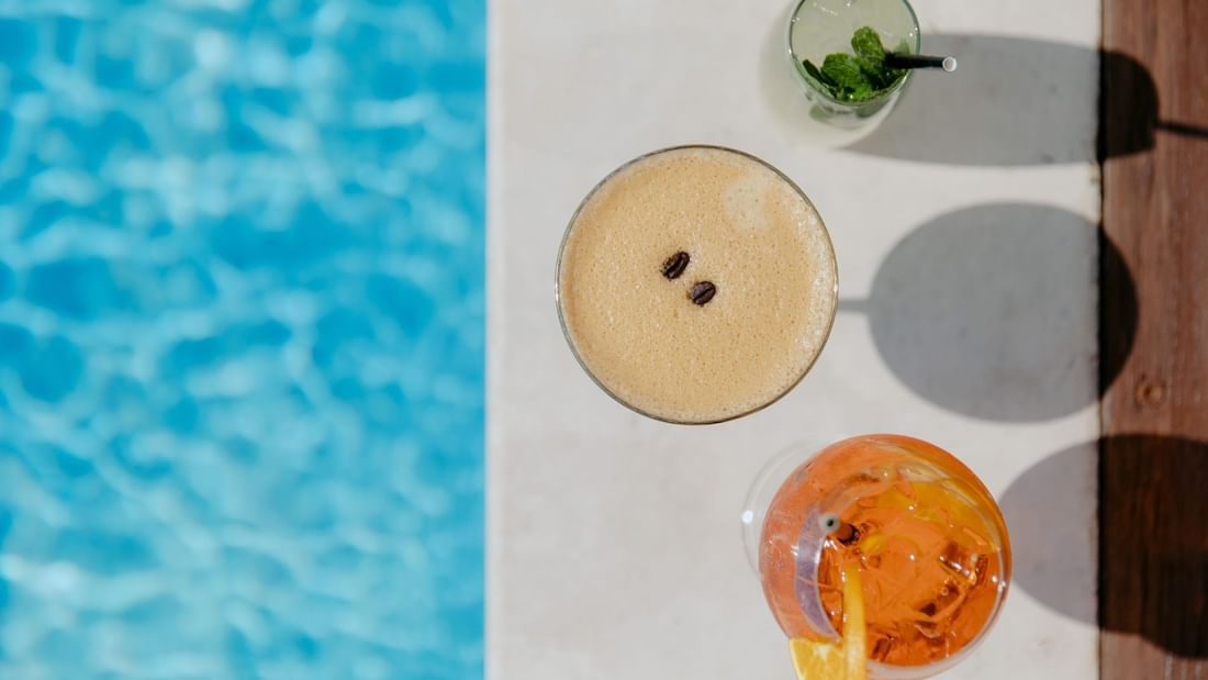 Three drinks by a pool, one with coffee beans, another with mint, and one with orange slice.