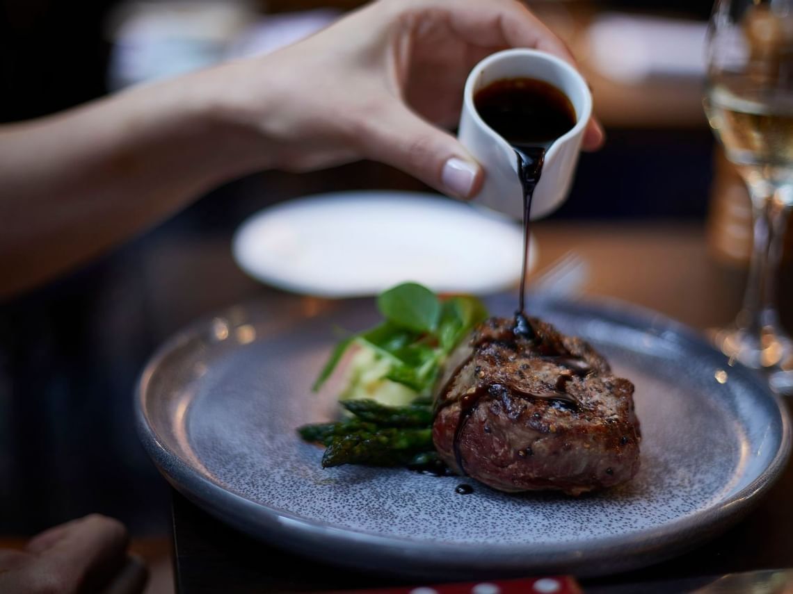 Hand pouring dark sauce onto a grilled steak with greens in The Reserve Restaurant at Amora Herencia Riverwalk Melbourne