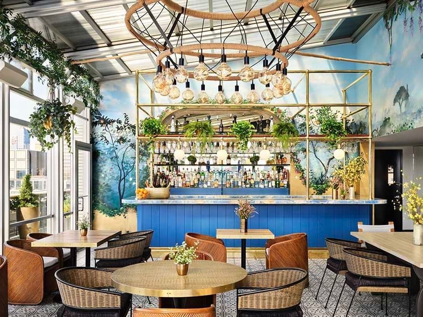 A restaurant with tables, chairs, a bar, plants, and a circular light fixture.