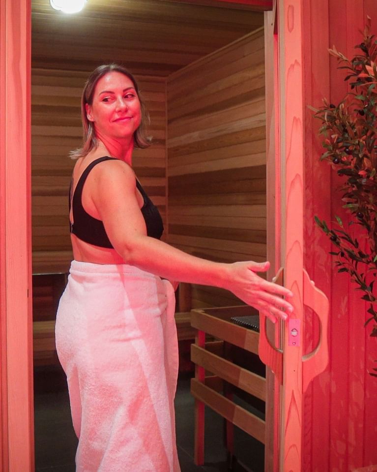 Woman in black top and white towel entering wooden sauna with red lighting 