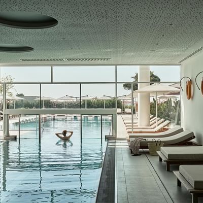 Innenbereich eines modernen Schwimmbads mit einer Person im Wasser und Liegen am Rand.