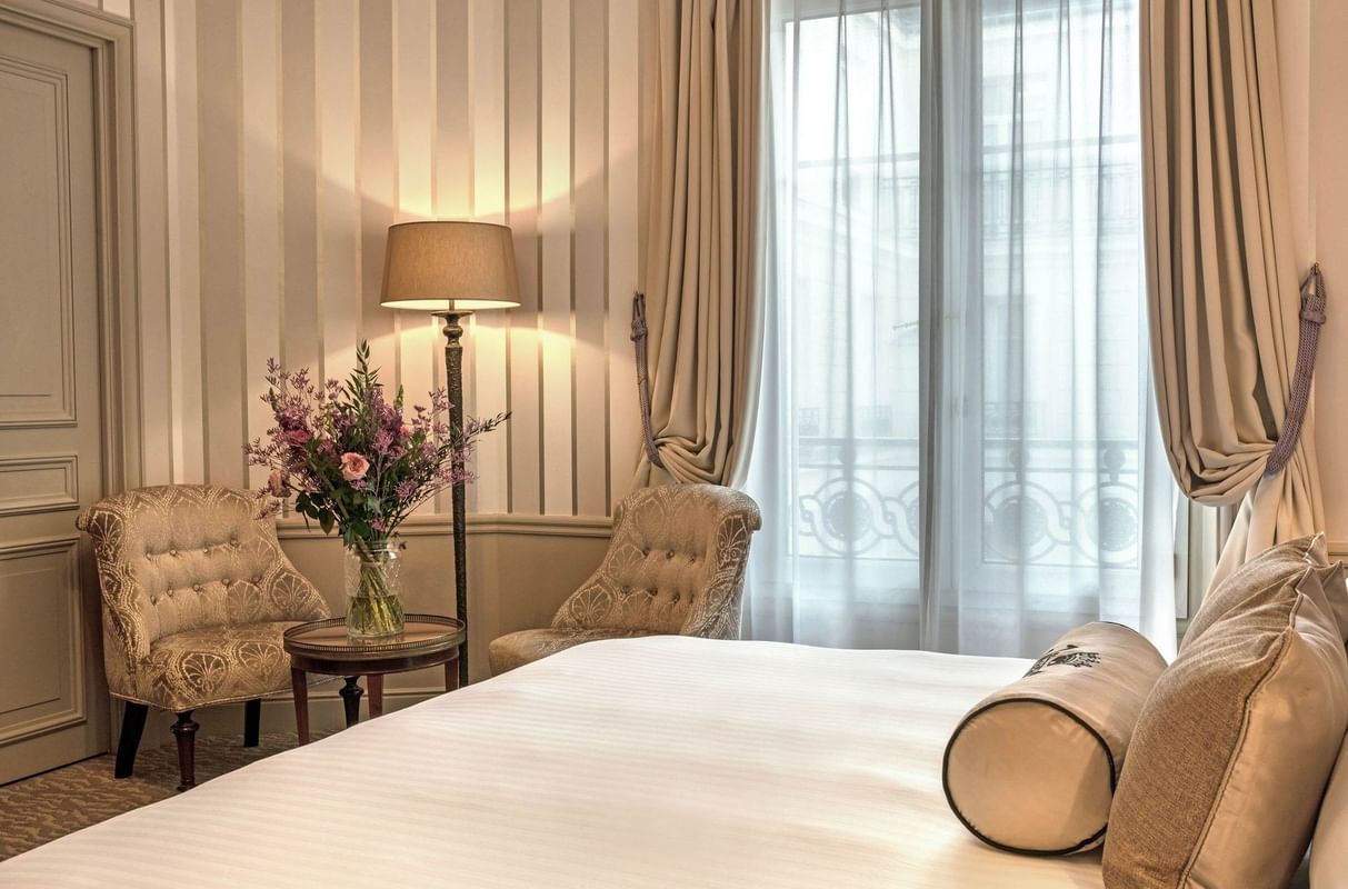 Chambre élégante avec lit double, fauteuils brodés, lampe allumée et grandes fenêtres à Hotel Westminster Warwick Paris