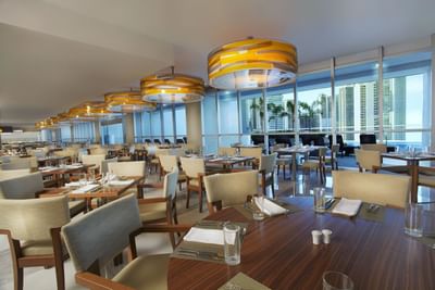 Bazar with wooden tables & cream chairs beneath pendant lights by tall windows overlooking the city at Megapolis Hotel Panama