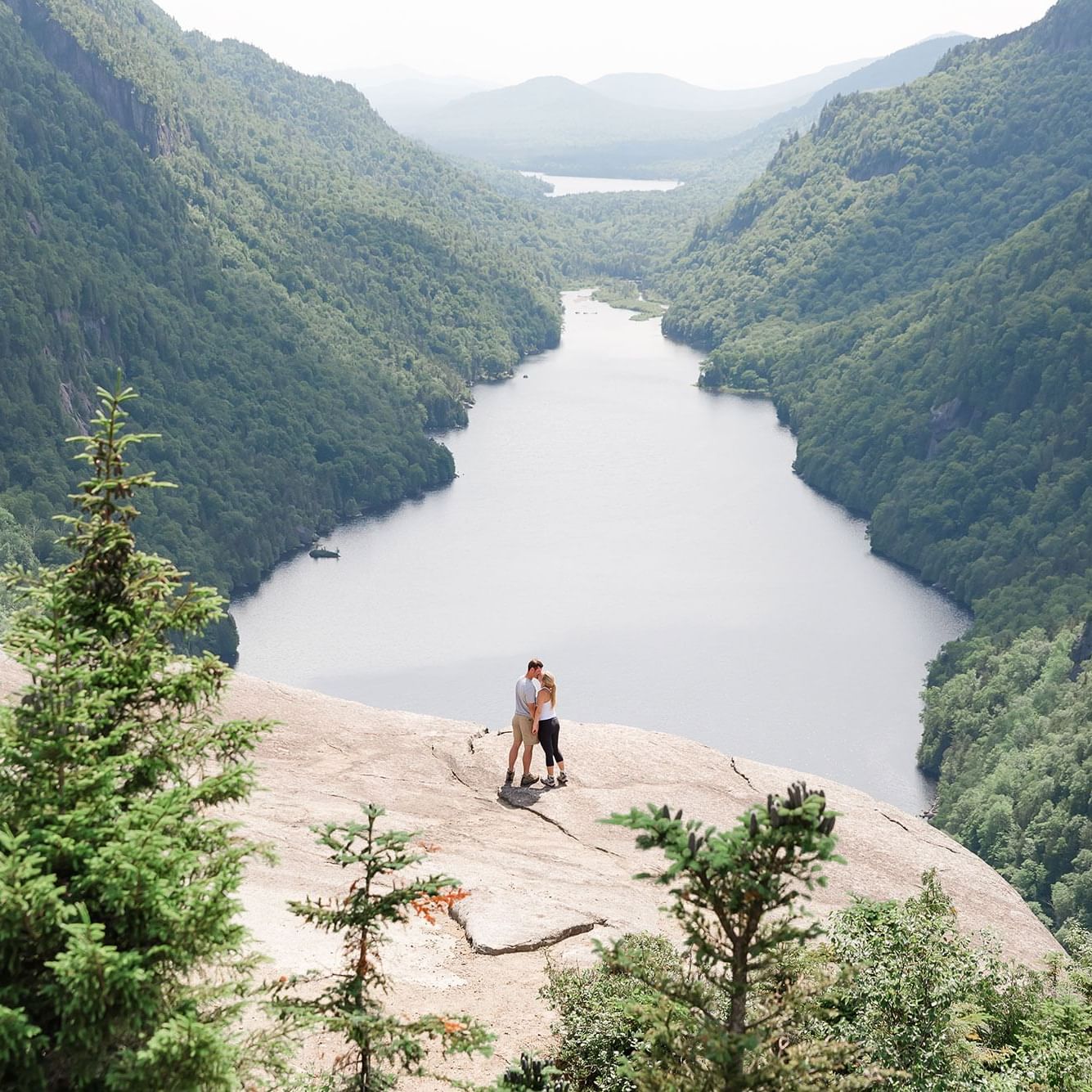 Couple standing on a rock overlooking a valley with a river and dense forest.