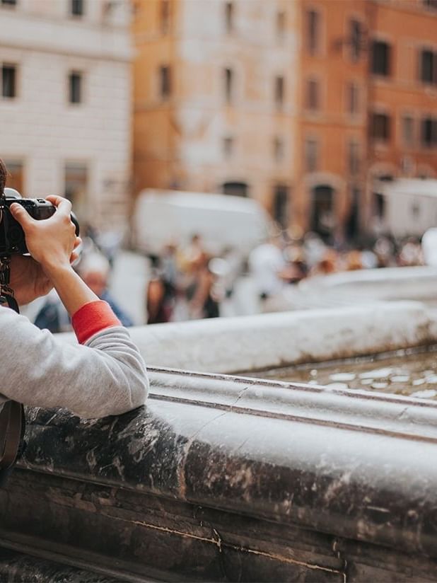 Person with a camera stands near a fountain, capturing the busy street near AG Hotels