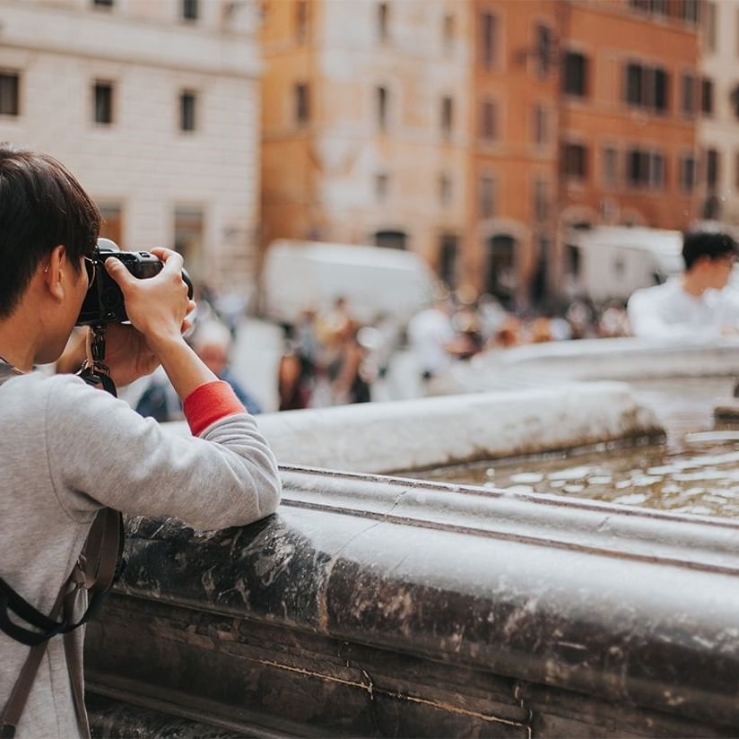 Person with a camera stands near a fountain, capturing the busy street near AG Hotels