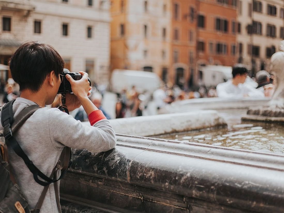 Person with a camera stands near a fountain, capturing the busy street near AG Hotels