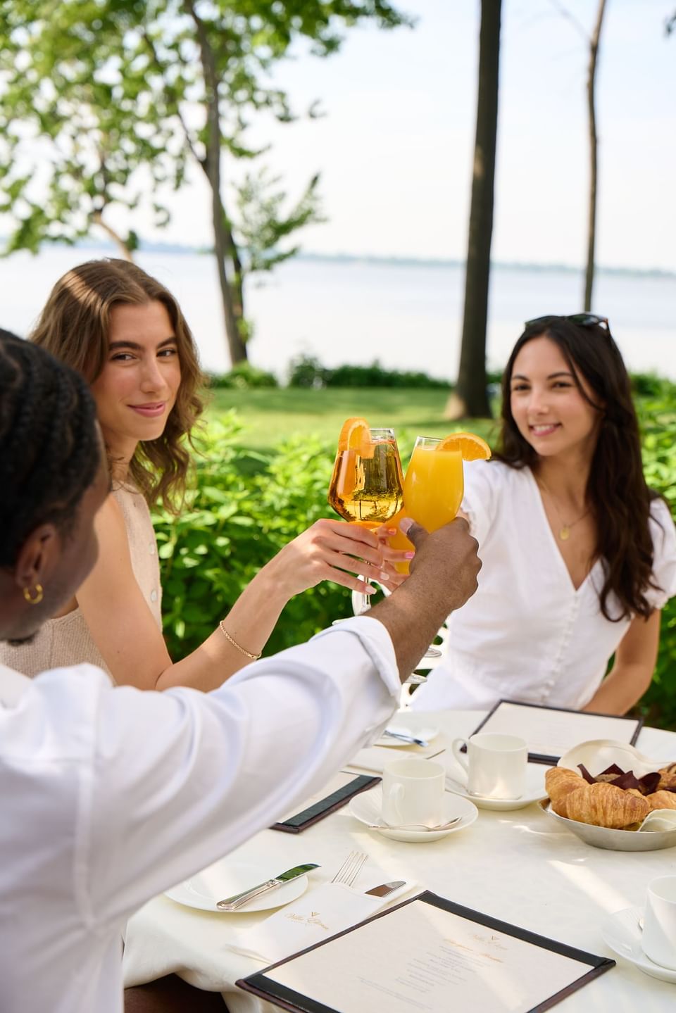 Friends toast with drinks at an outdoor table overlooking a serene lake by Chateau Vaudreuil