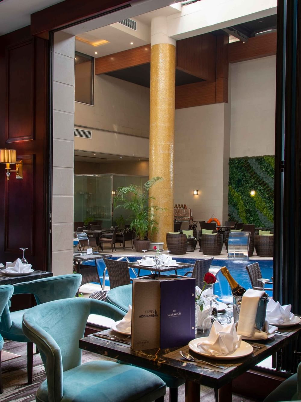 Teal velvet chairs set dining tables overlooking a pool and indoor garden atrium in Cigar Lounge at Warwick Al Khobar