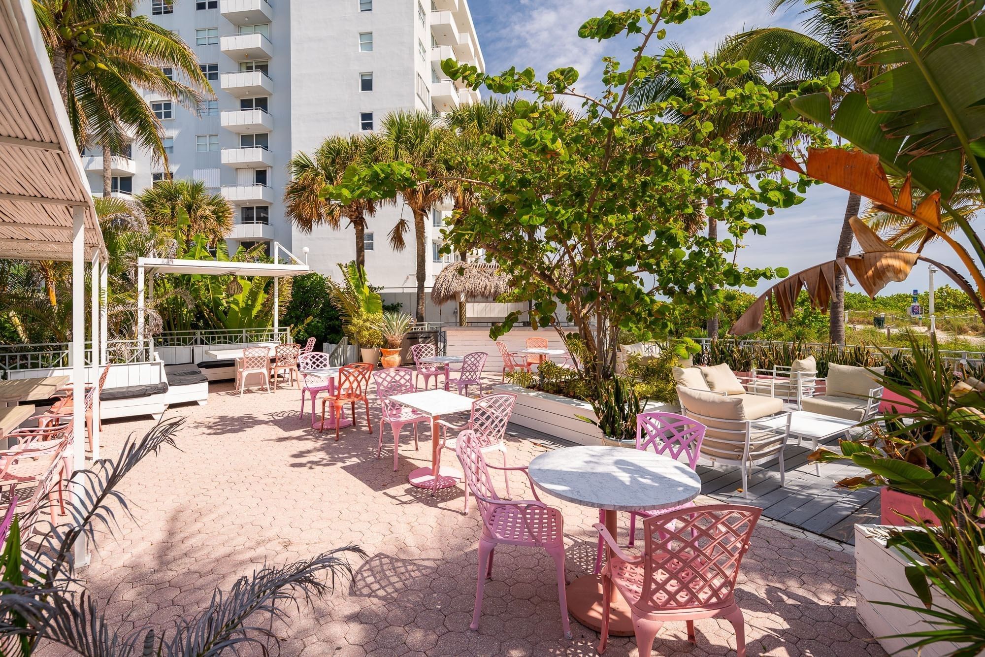 A sunny outdoor seating area with pink metal chairs and white tables surrounded by trees and potted plants.