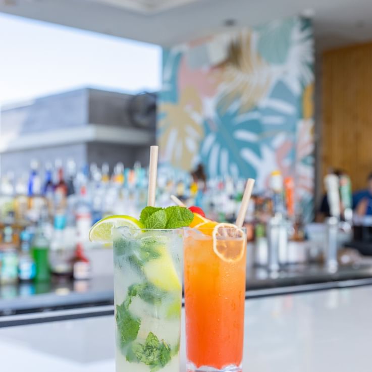 Two colorful cocktails on a bar with a tropical background