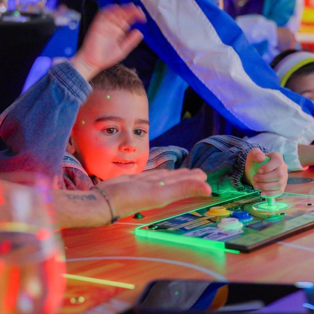 Child gripping a glowing green joystick on an arcade console, surrounded by colorful lights at Hotel X Toronto