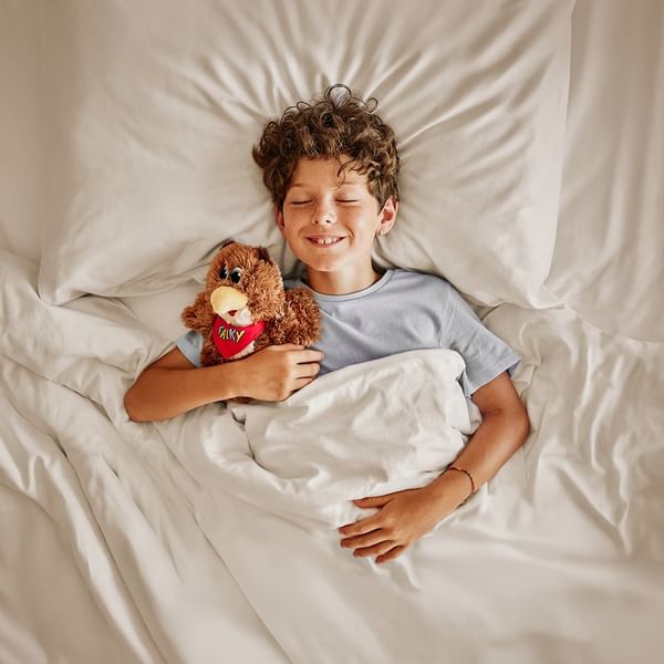 A boy lies in bed with a stuffed toy, smiling with eyes closed, under white bedding.