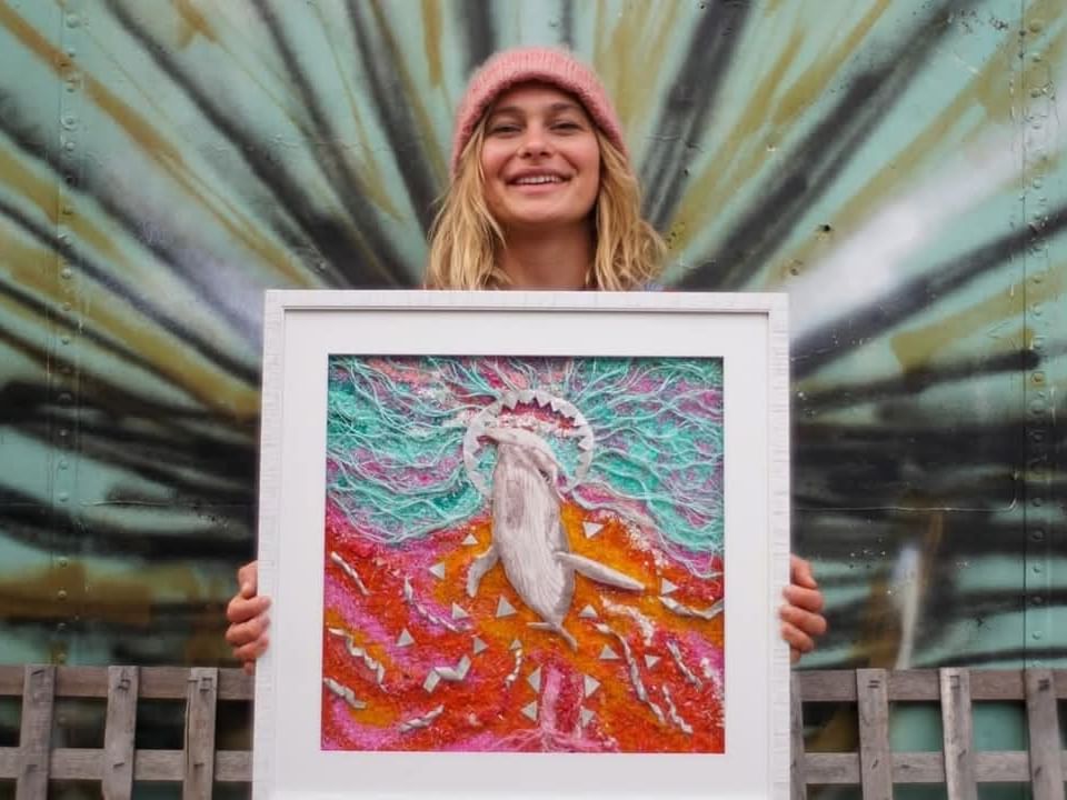 Woman in pink beanie smiling while holding colorful framed artwork in front of painted corrugated metal backdrop.