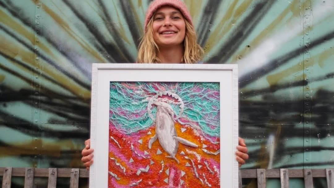 Woman in pink beanie smiling while holding colorful framed artwork in front of painted corrugated metal backdrop.