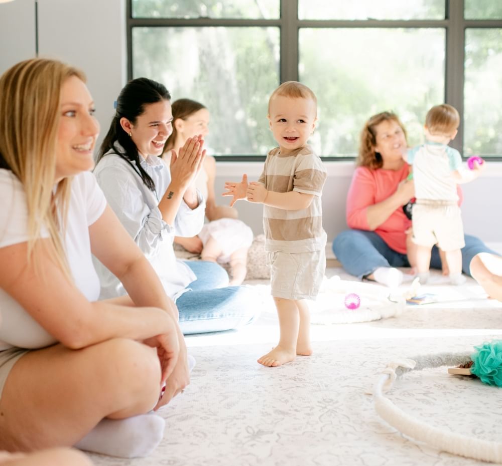 A toddler boy surrounded by moms in a circle claps and smiles, while another child plays in the background.