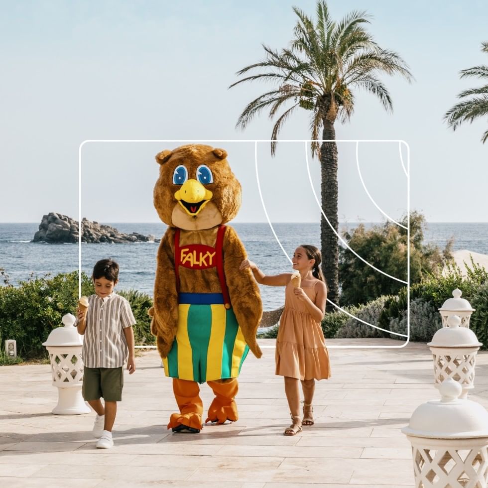 Zwei Kinder halten die Hände des Maskottchens Falky vor Meerblick und Palmen im Falkensteiner Resort Capo Boi.