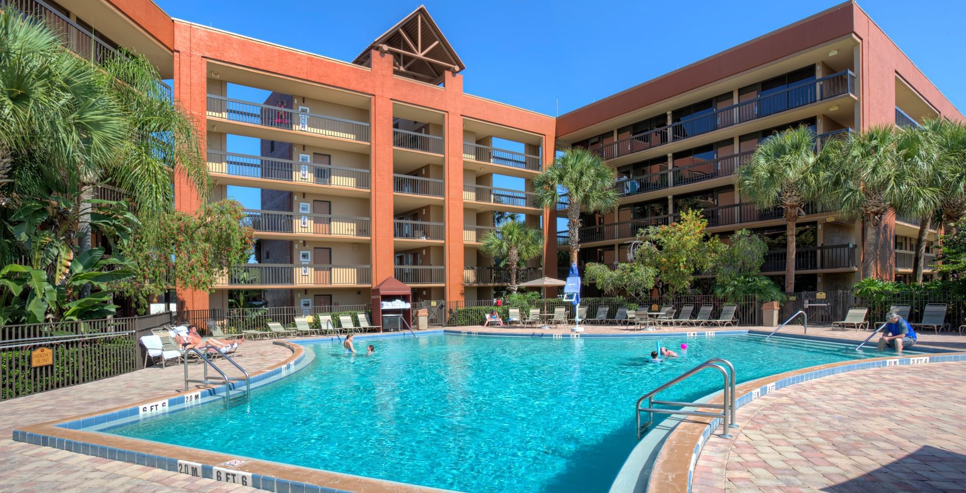 A large outdoor hotel surrounding a beautiful pool with palm trees and lounge chairs. 