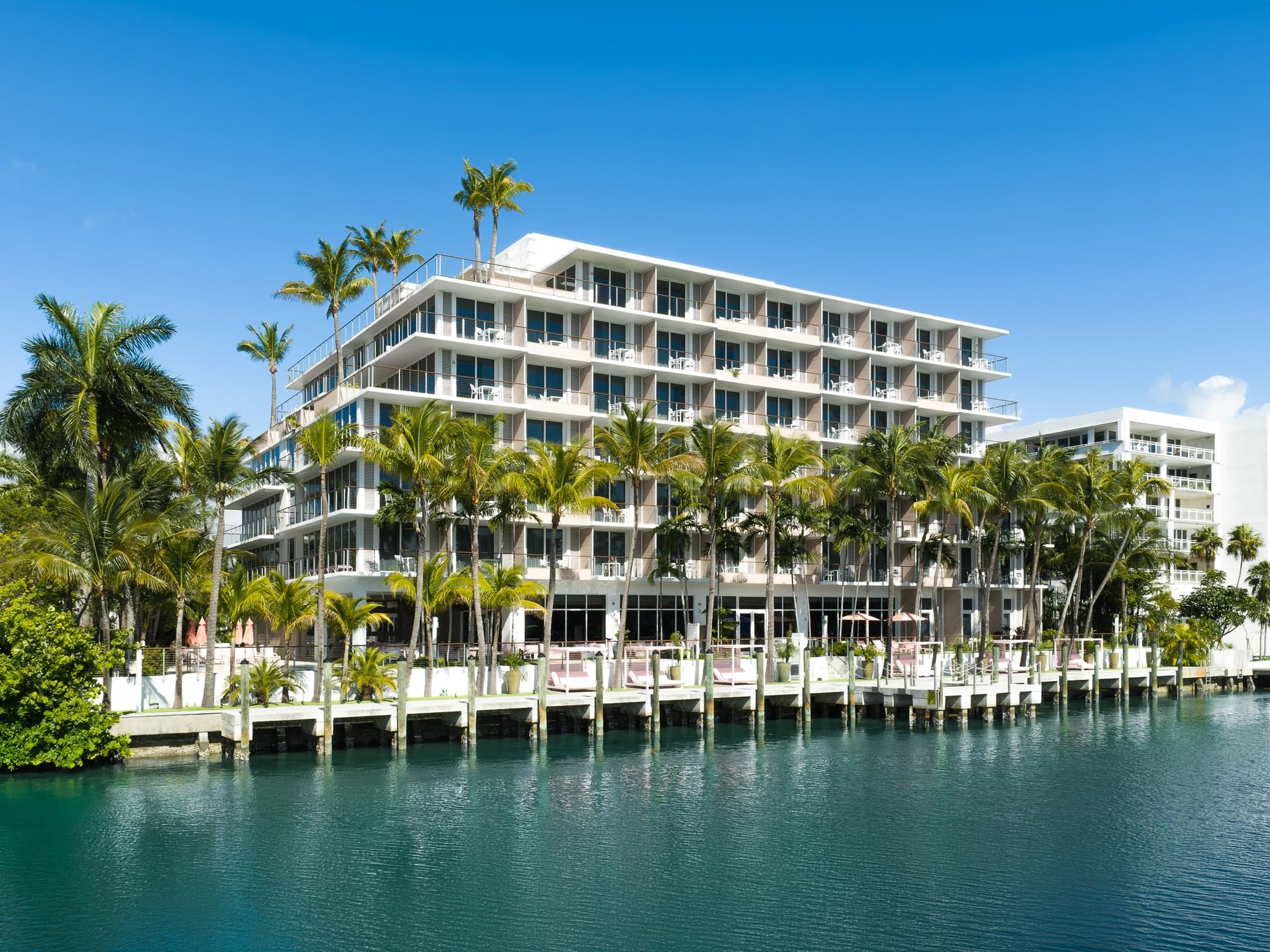 A large white building with balconies and palm trees in front of it, overlooking a body of water.
