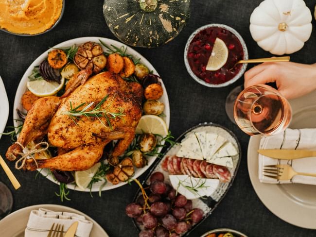 Manos sosteniendo copa de vino sobre mesa con pavo asado, guarniciones y postres, listos para una comida festiva.