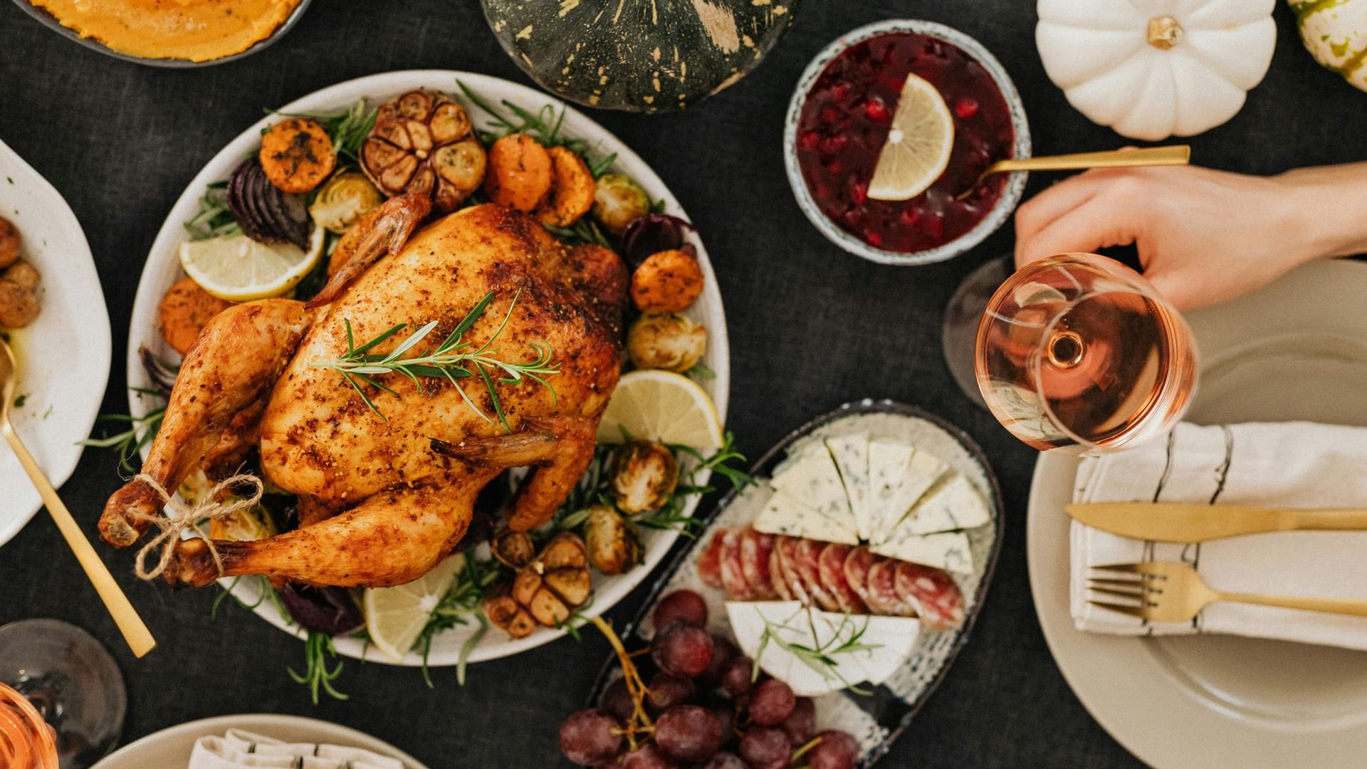 Manos sosteniendo copa de vino sobre mesa con pavo asado, guarniciones y postres, listos para una comida festiva.