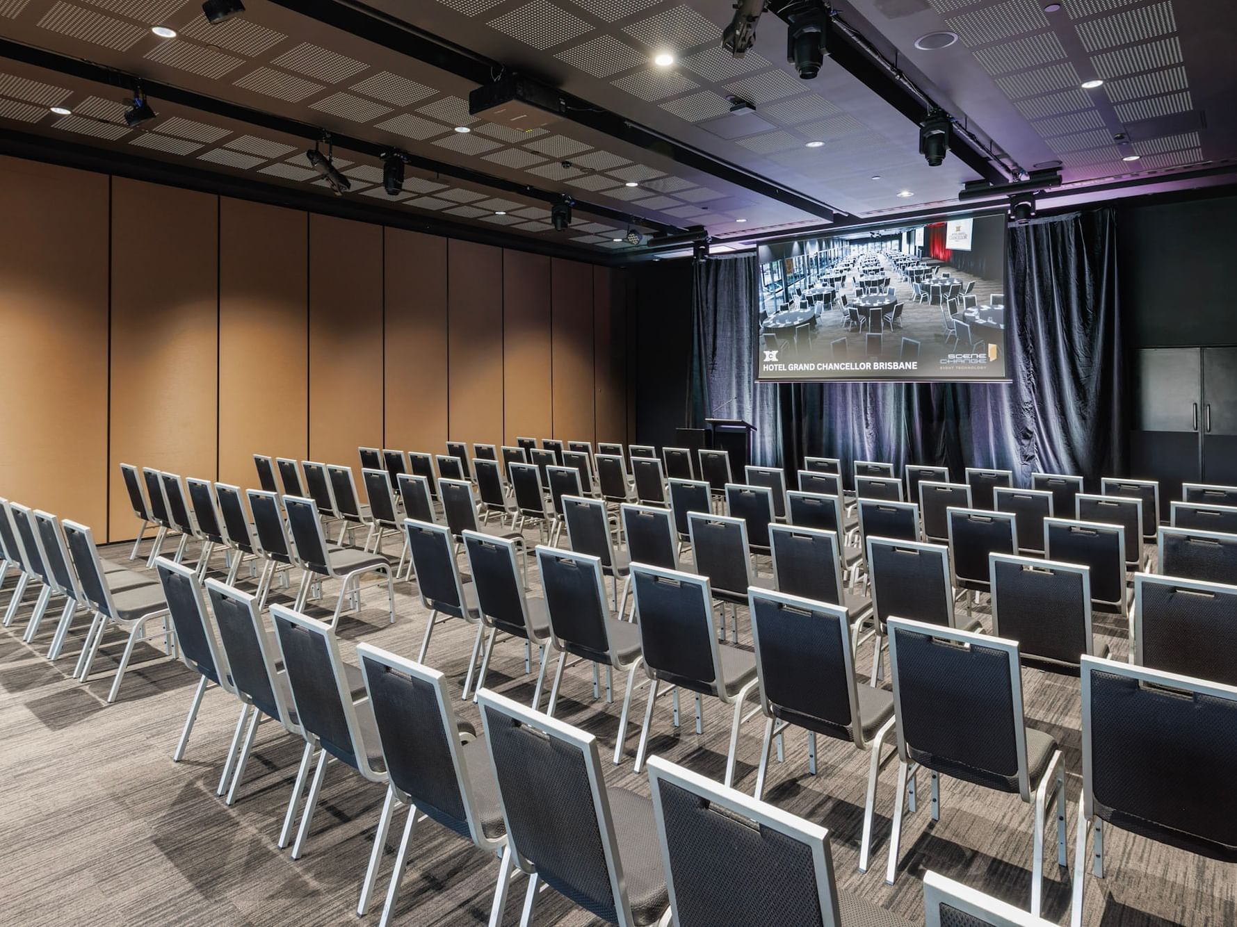 A large, modern conference room at Hotel Grand Chancellor Brisbane set up in theater style with rows of grey chairs, professional lighting, and a large projector screen.