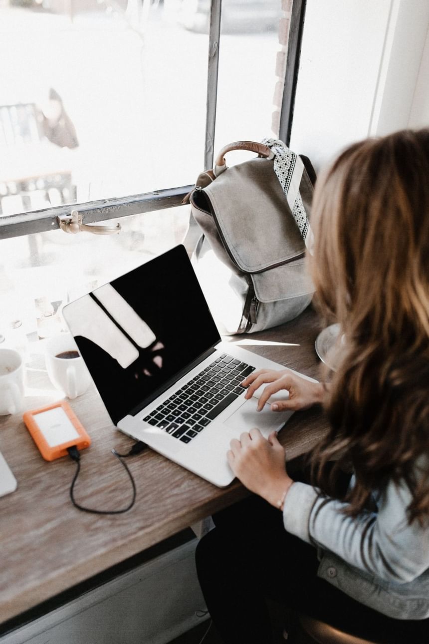 Woman using a laptop by a coffee and a backpack on a wooden counter beside a bright window at the Hotel Saint Clair