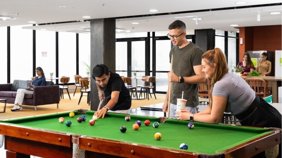 Three people play pool in a lounge with a green table, and others sit nearby.