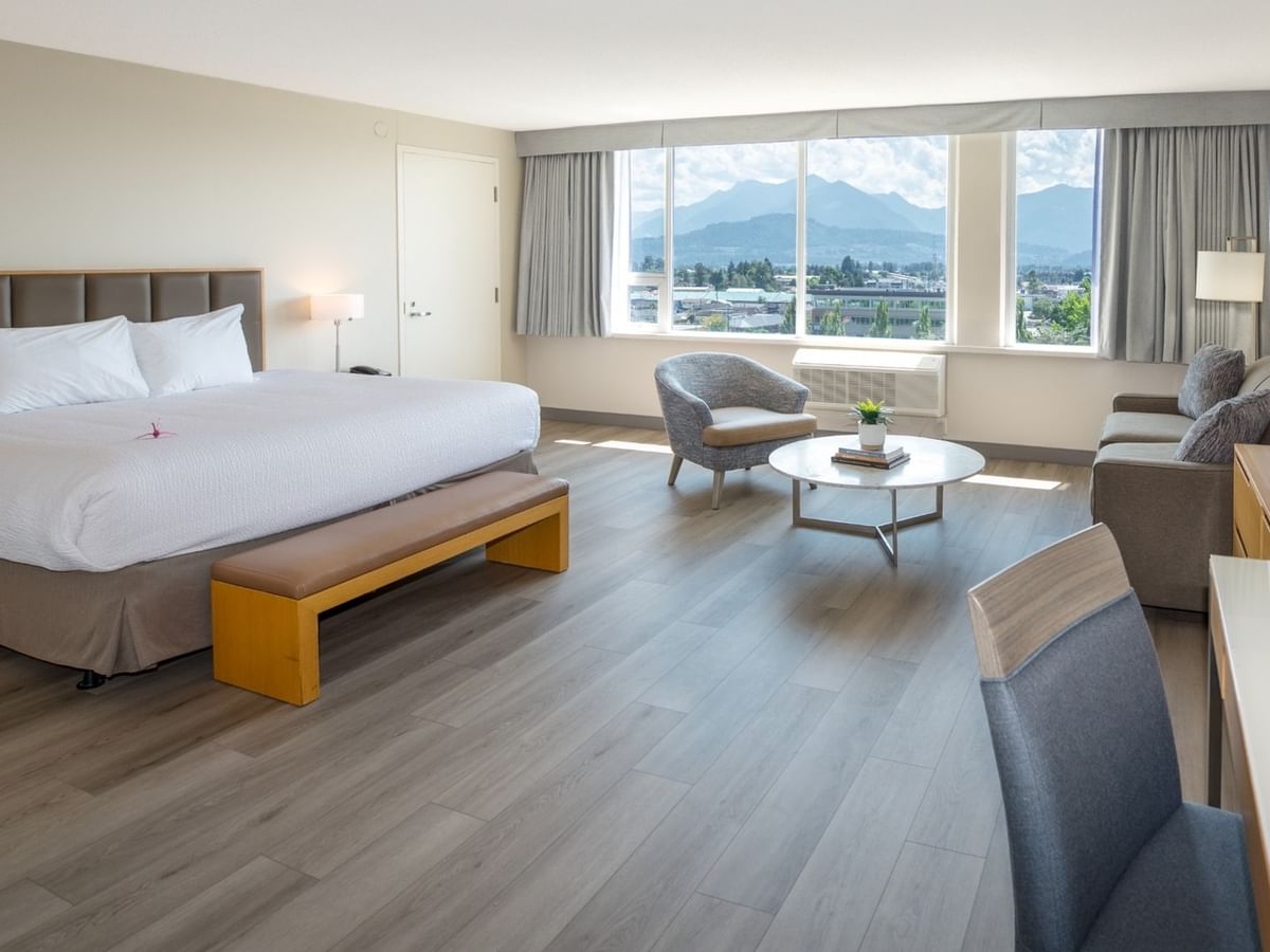 Hotel room with large bed, seating area, desk, and mountain view from large windows.
