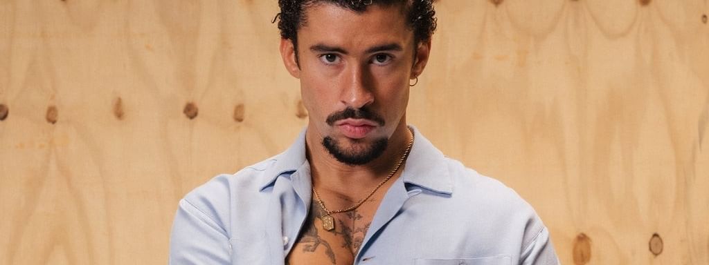 Bad Bunny with curly hair and tattoos on his chest stands against a wooden wall, wearing a light blue shirt.