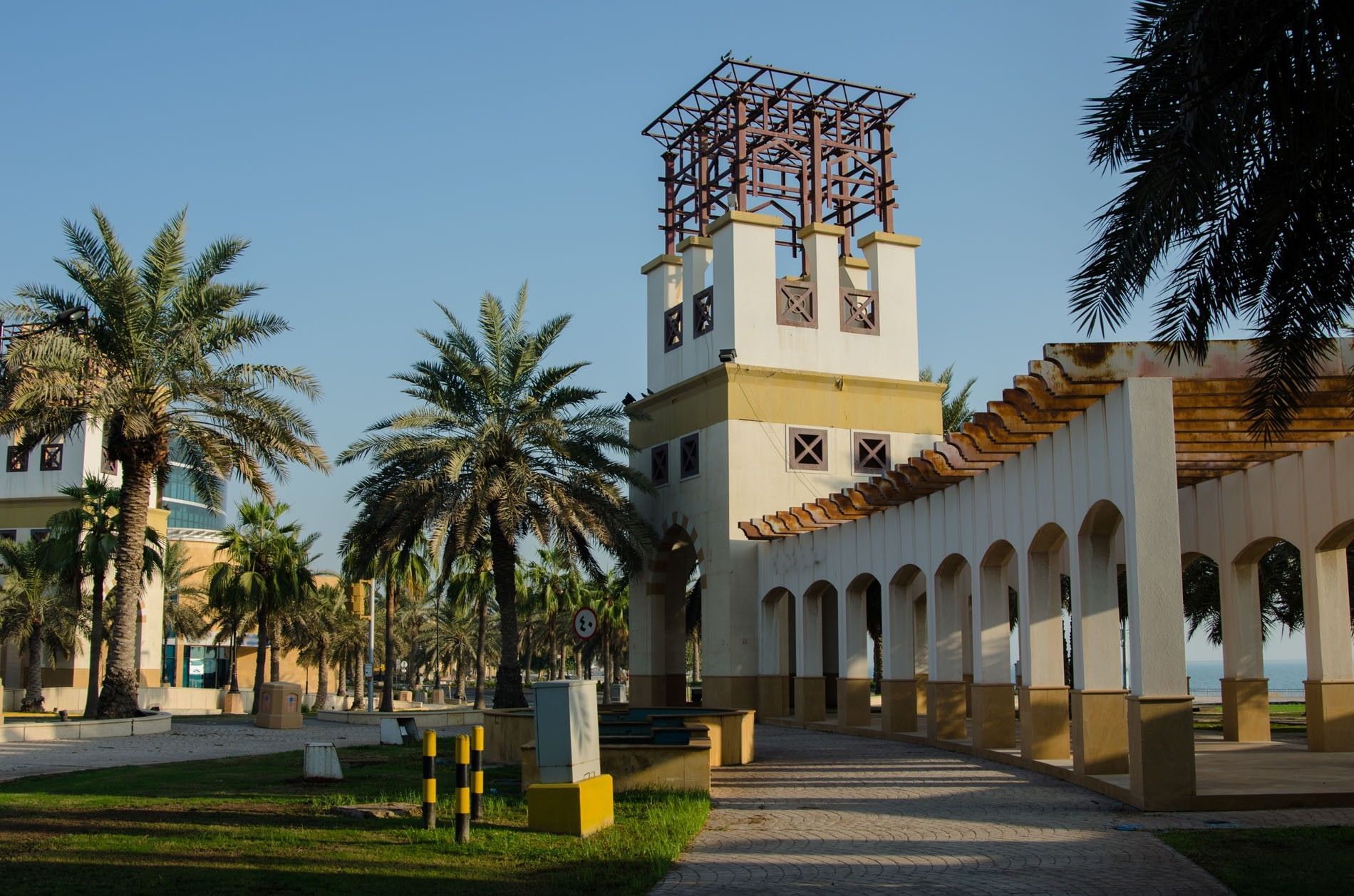 Stone walkway by a white arched structure and palm trees near a park at Warwick Al Jubail
