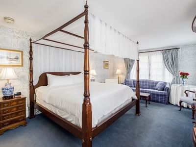 Four-poster bed under white canopy by a plaid loveseat and round table in Deluxe King with Sofabed at Simsbury 1820 House