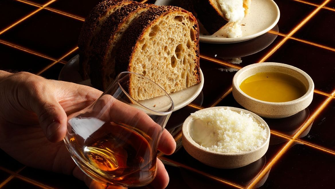 A hand holds a glass of amber liquid next to bread slices, olive oil, and cheese.