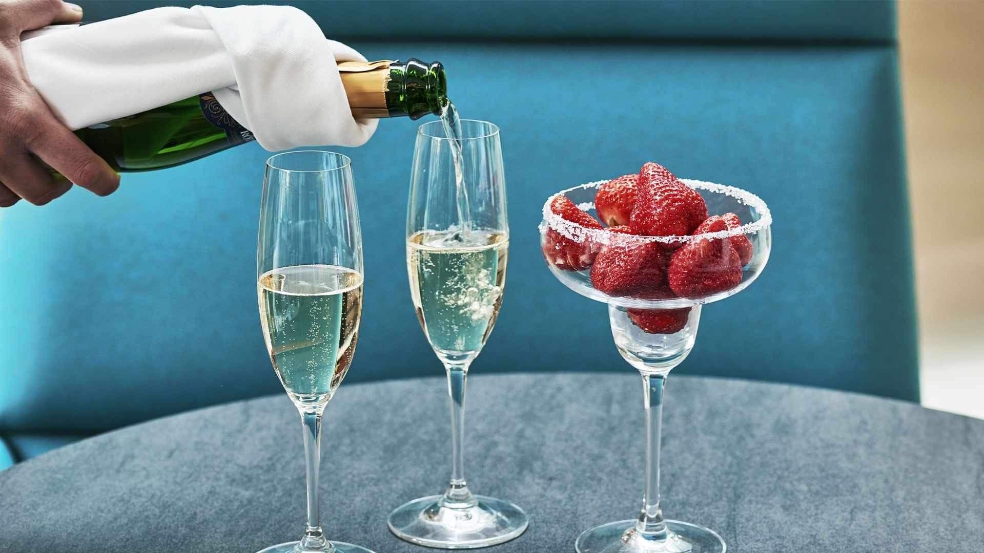 Hand gießt Champagner in zwei Gläser, daneben eine Schüssel mit Erdbeeren.