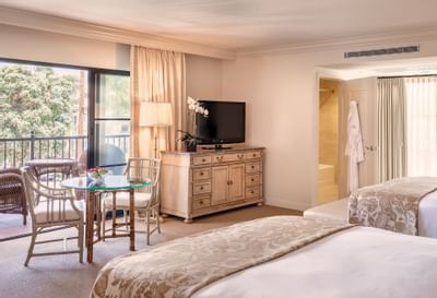 Standard Two Queen at Santa Barbara Inn with two beds, a TV, and a balcony with a dining area overlooking lush greenery.