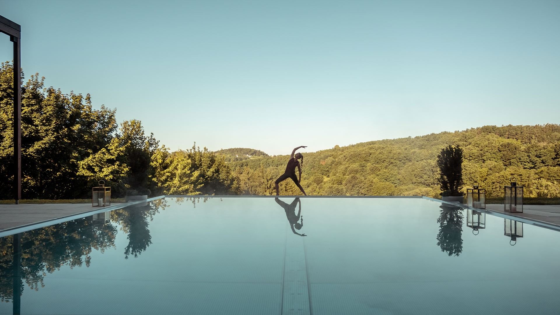 Person doing yoga pose on pool deck with reflection in water at Falkensteiner Group