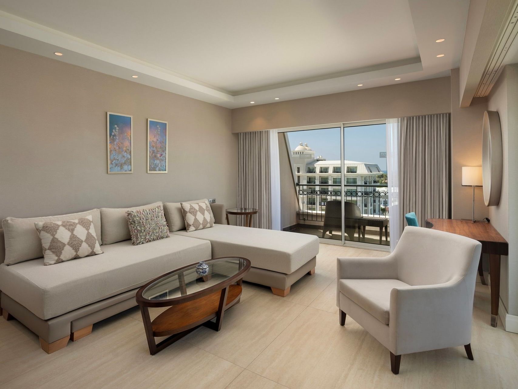 Modern living room with beige sectional sofa, armchair, glass coffee table, and balcony overlooking resort buildings.