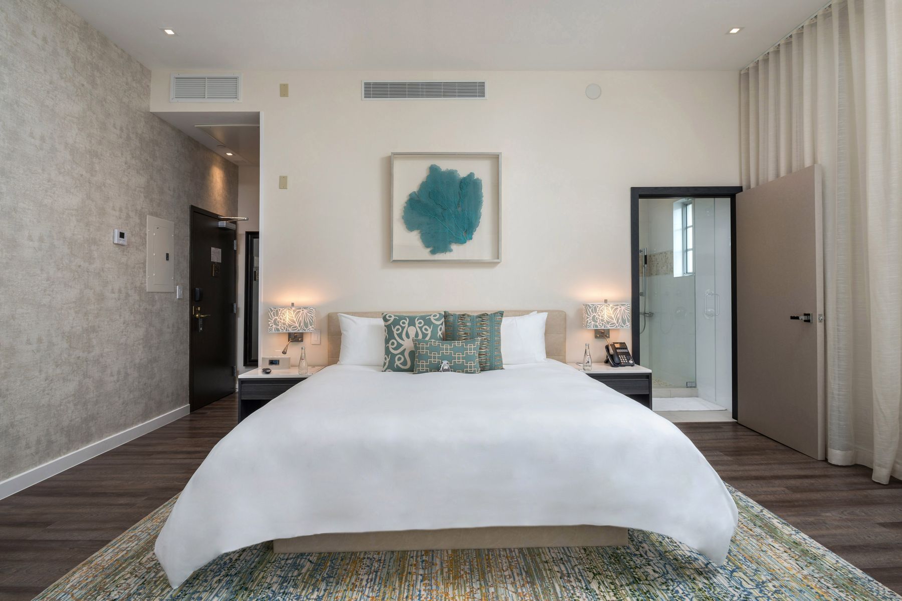 A large bed with white linens and teal pillows is centered in a room with a shower and doorway to the right.