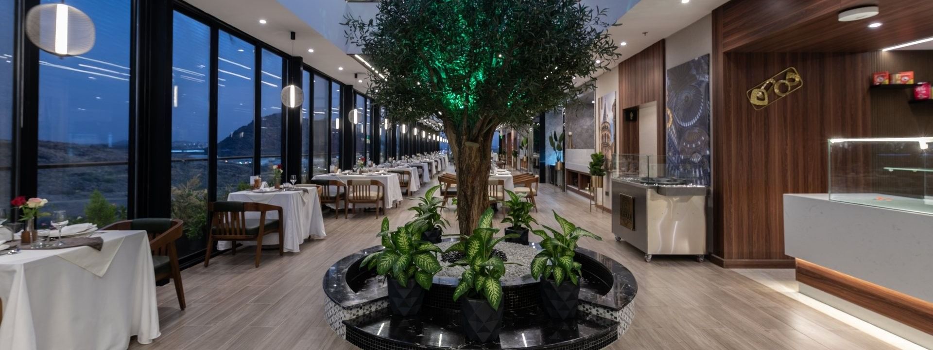 Large tree in a marble planter by dining tables under a blue glass skylight in Karkan Restaurant at Warwick Al Taif Hotel