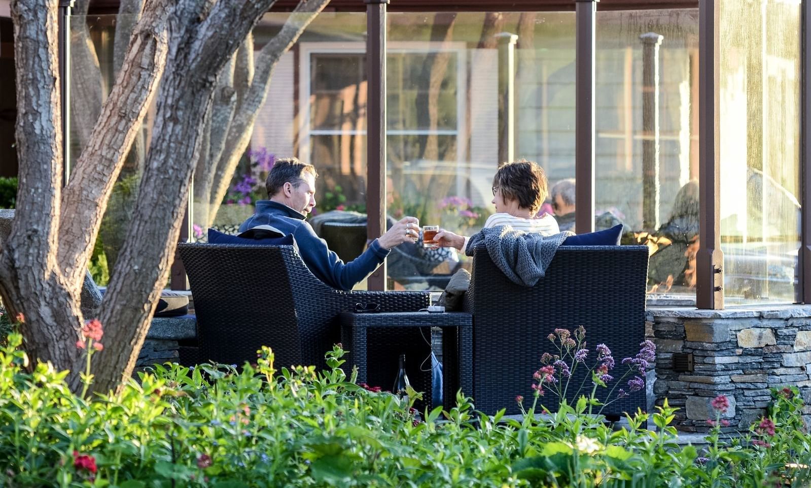 A couple toasts with drinks at an outdoor table with a glass structure in the background.