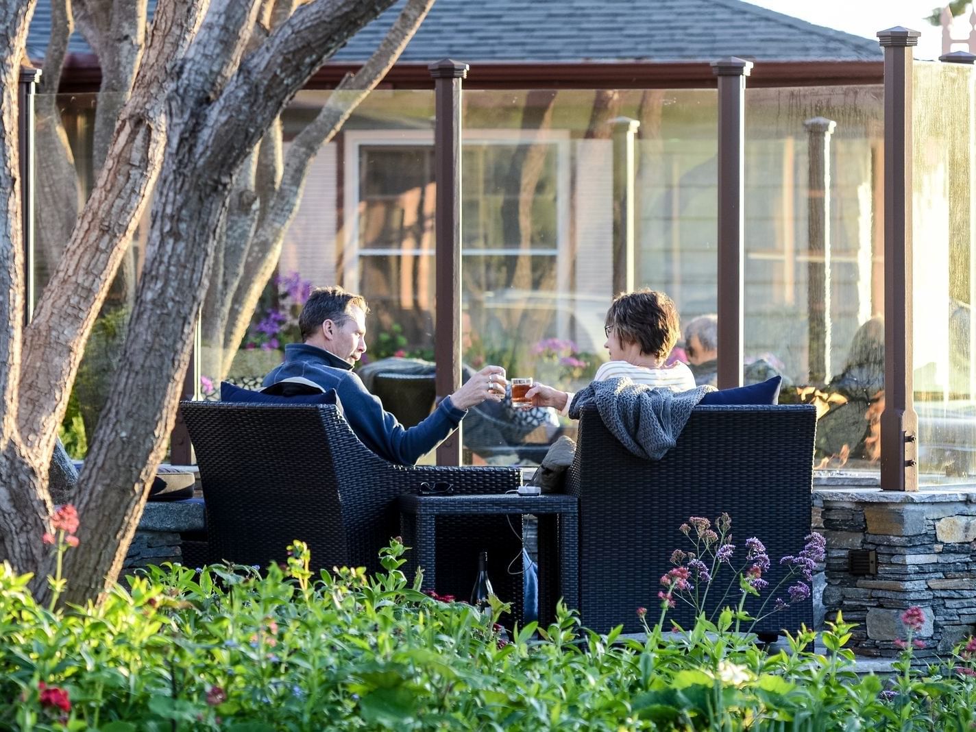 Two guests in wicker chairs by a glass fire pit under a shady tree at one of the Beachfront hotels in Cambria, Sea Otter Inn