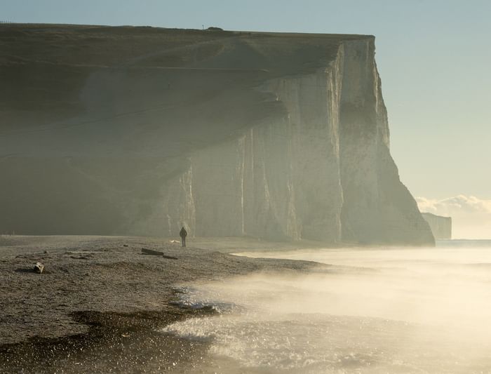Person stands on a beach before a cliff with a path on top, and the sea is in the foreground at Birling Gap