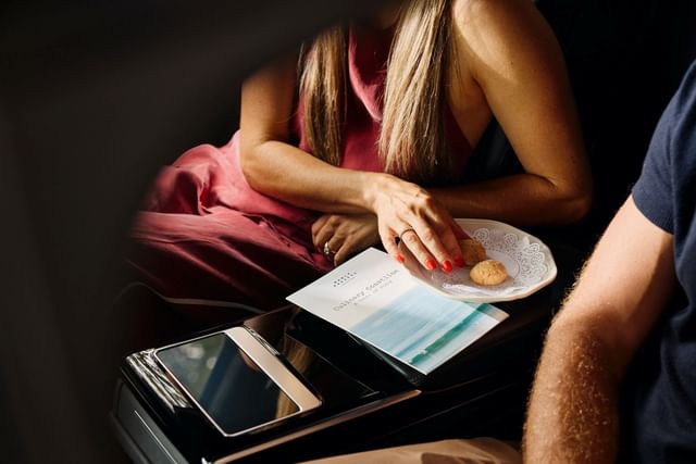 Elegant woman in a pink dress at Elysium Manly, reaching for sweet cookies by a menu book under sunlight in a dark car