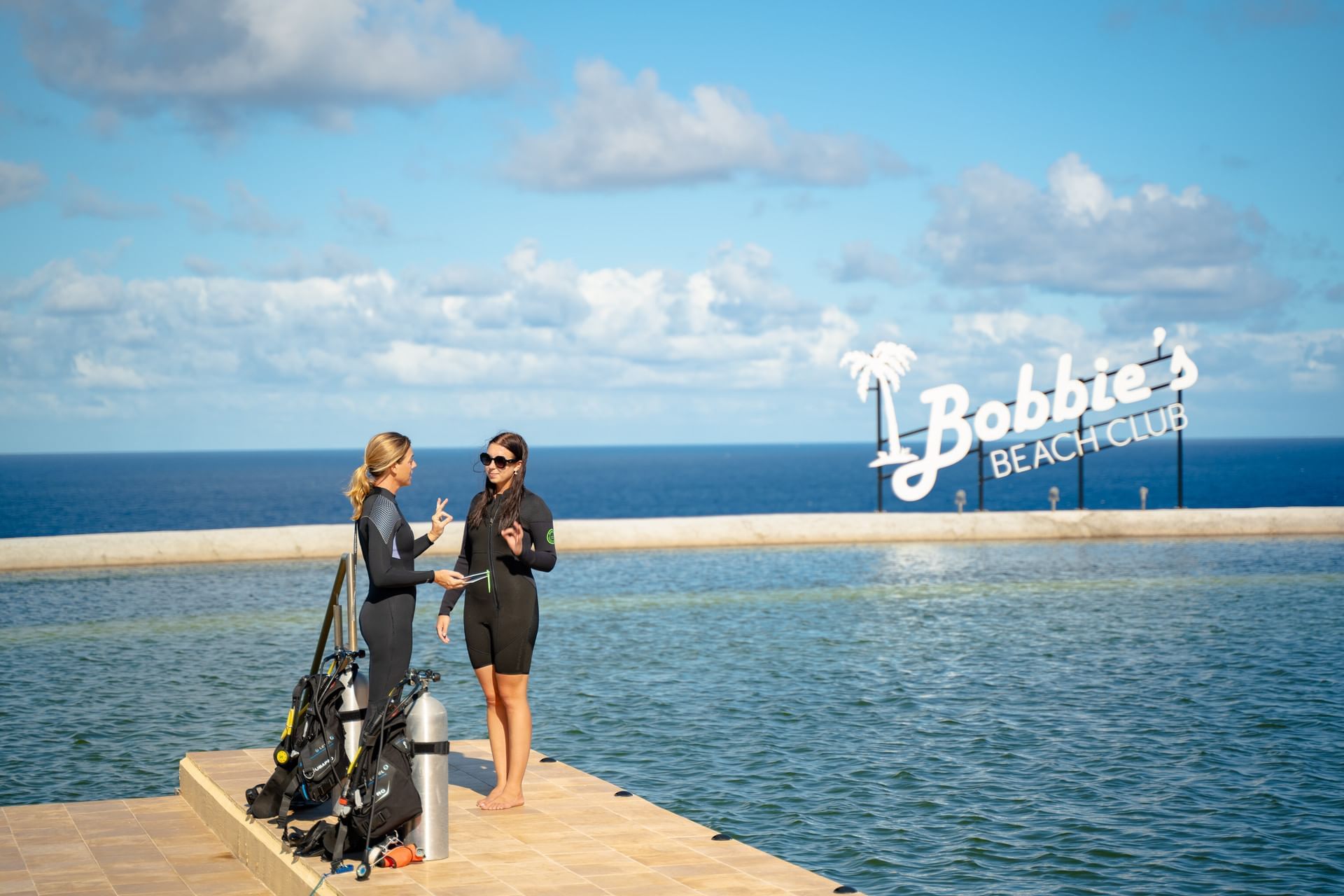 Twee duikers praten bij een zwembad met Bobbie's Beach Club-bord en uitzicht op de oceaan bij Golden Rock Resort