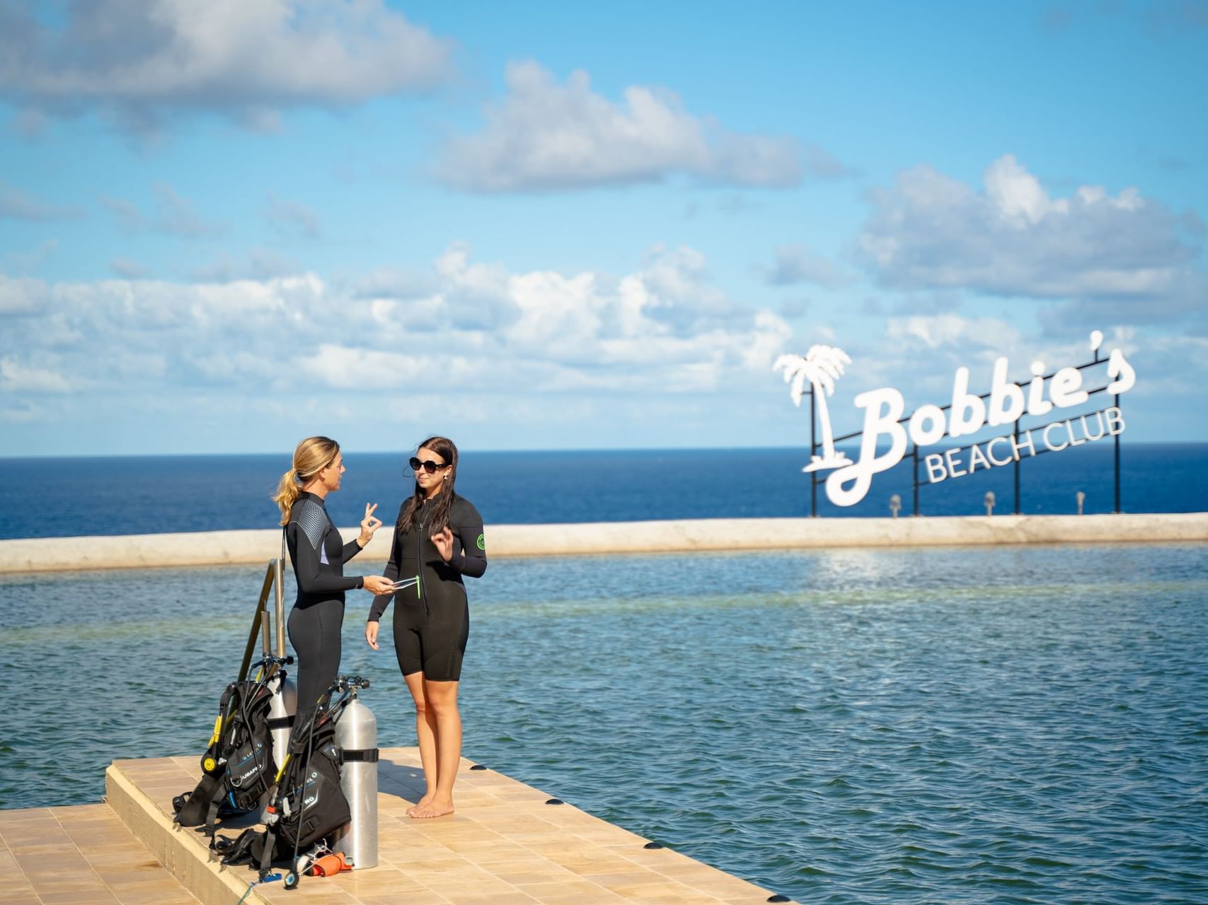 Two scuba divers talking by a pool with Bobbie's Beach Club sign and ocean view at Golden Rock Resort