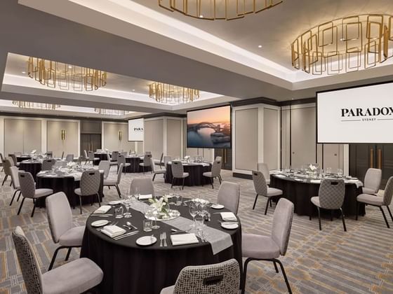 Banquet hall with round tables, chairs, and screens displaying PARADOX SYDNEY, ready for an event.